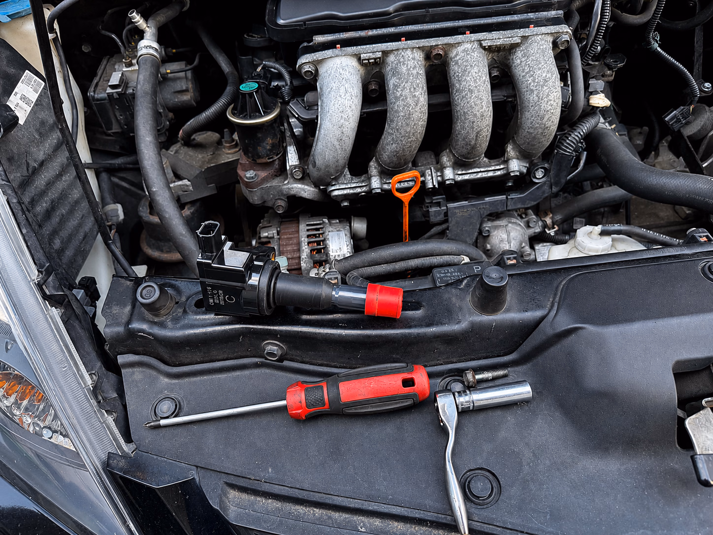 Car engine bay with tools including a screwdriver, ratchet wrench, and ignition coil on the engine cover.