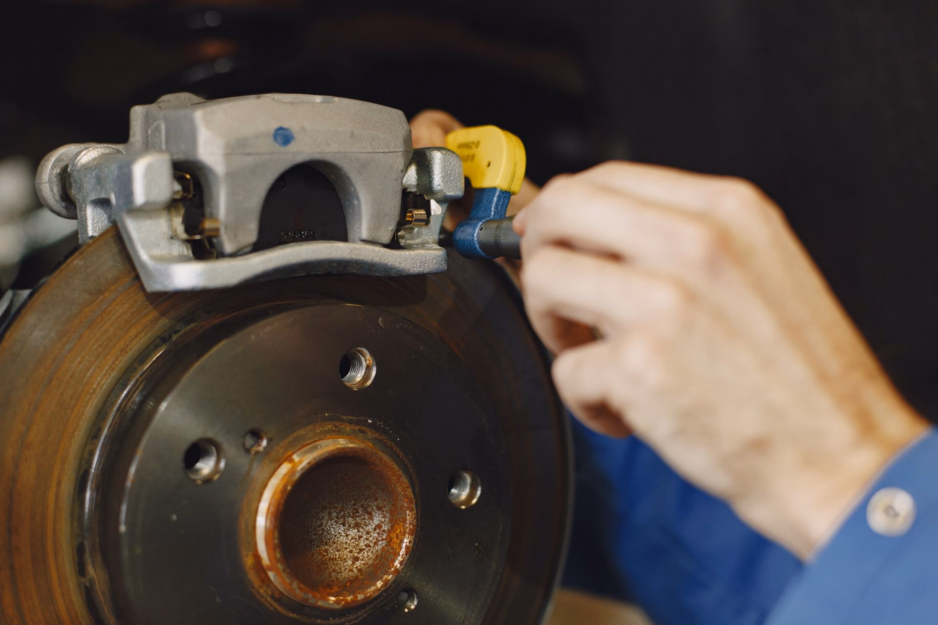 Hand using a micrometer to measure a car brake disc near its caliper.