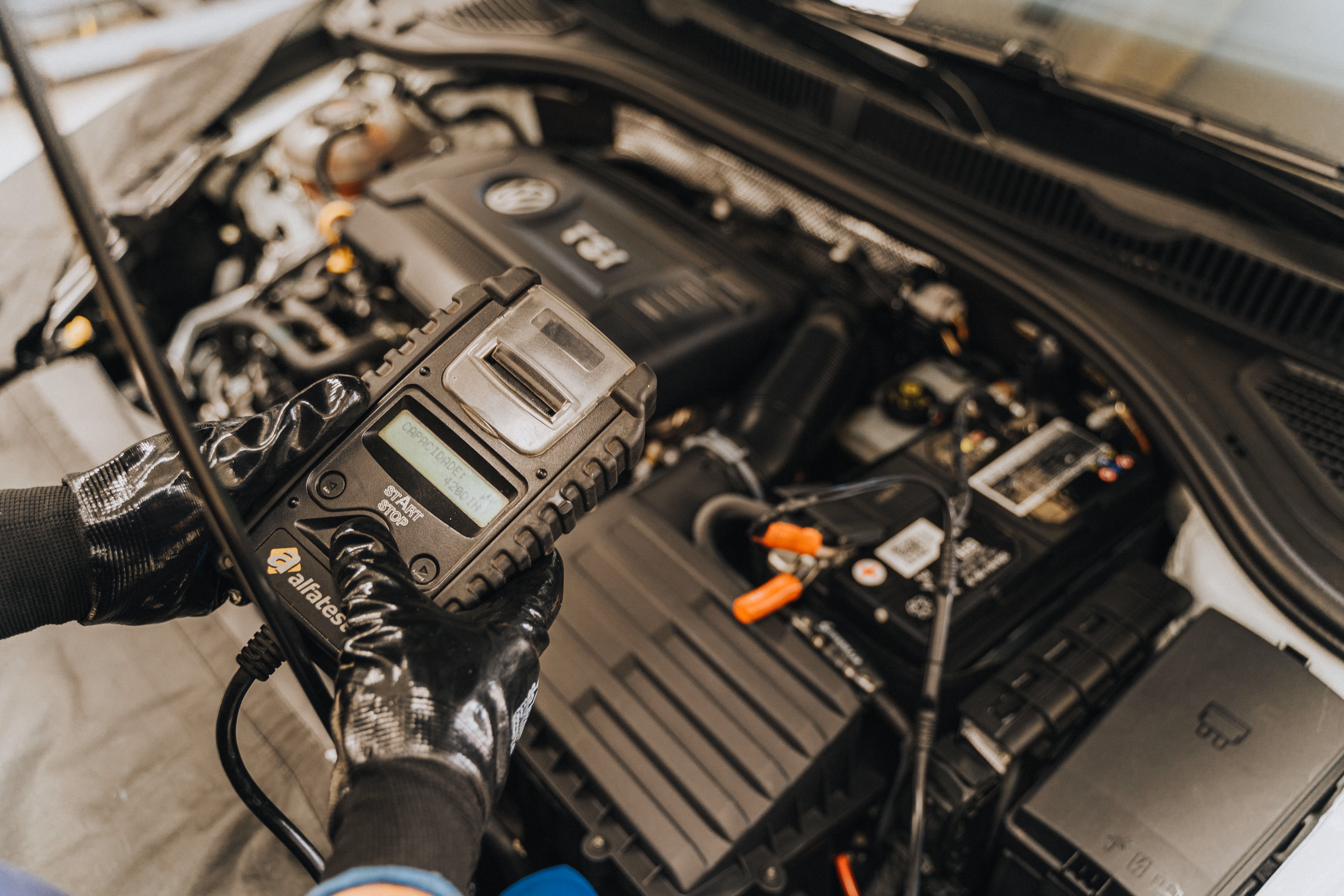 Gloved hands holding a battery tester reading capacity inside a car engine compartment.