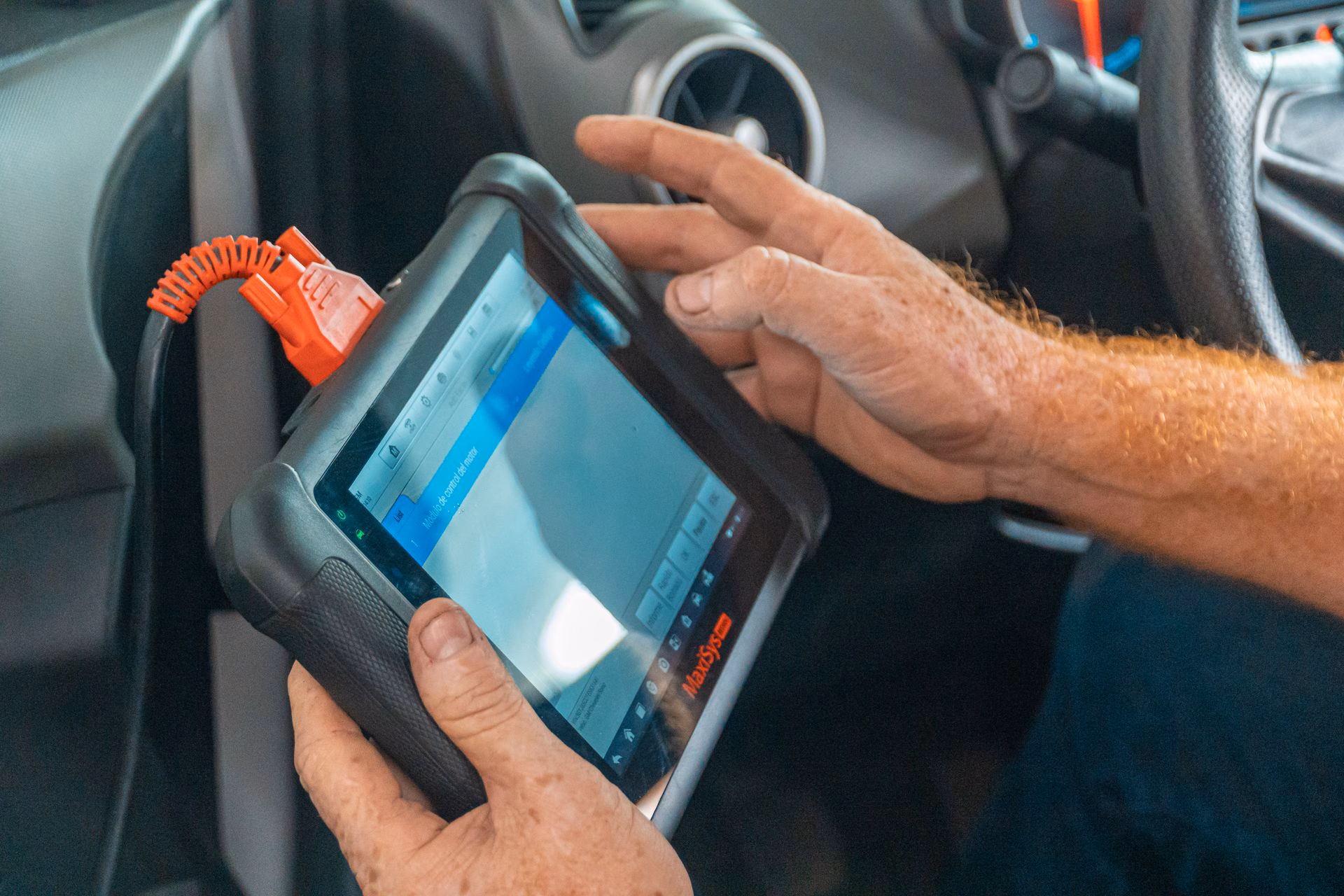 Person holding a MaxSys diagnostic tablet inside a vehicle, connected with an orange cable.