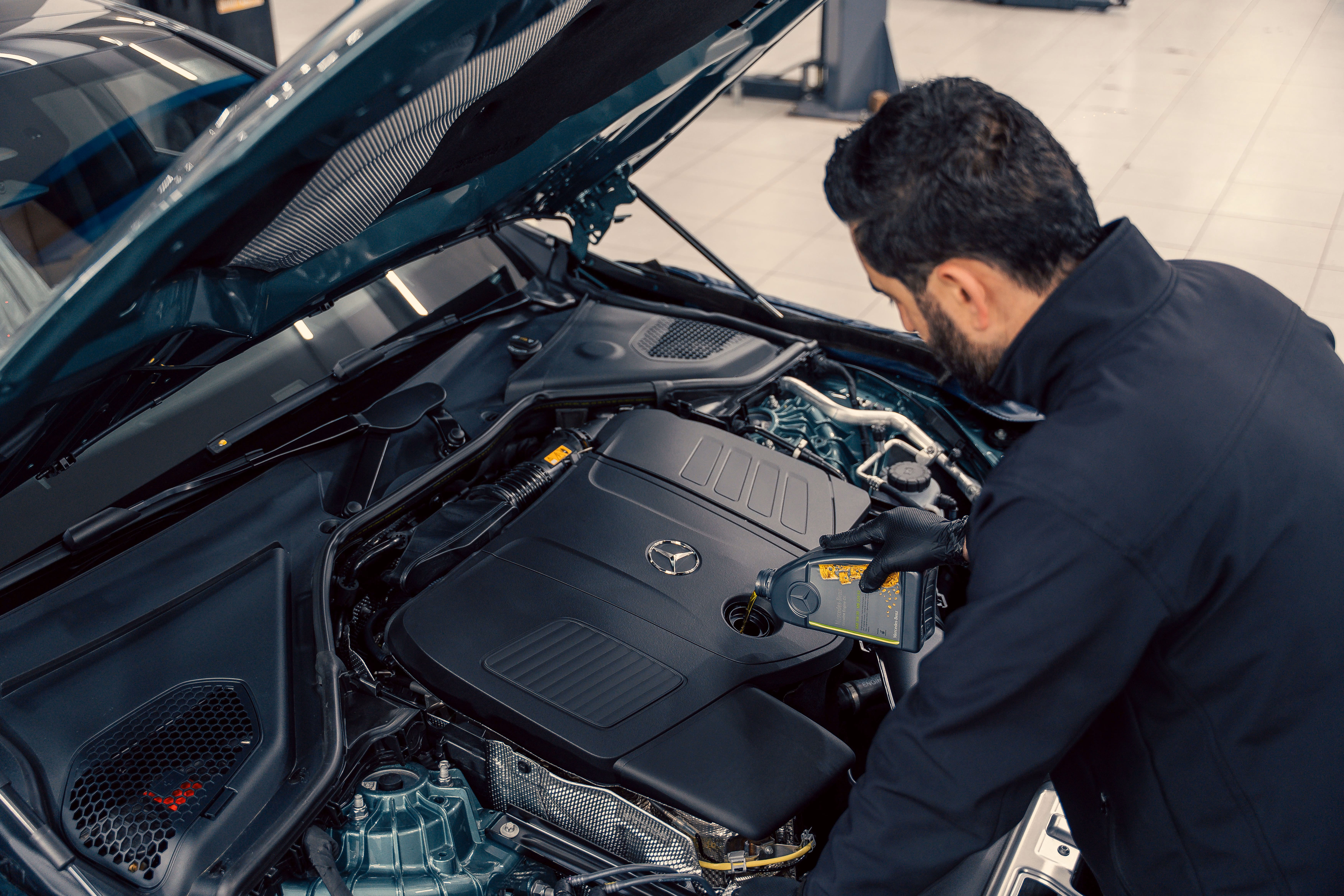 Mechanic pouring oil into a Mercedes car engine under an open hood in a garage.